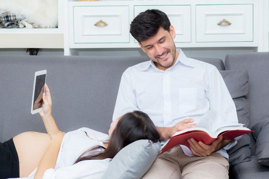 Asian couple family using a tablet at home, the young mother is pregnant lying on sofa looking tablet and husband reading book.