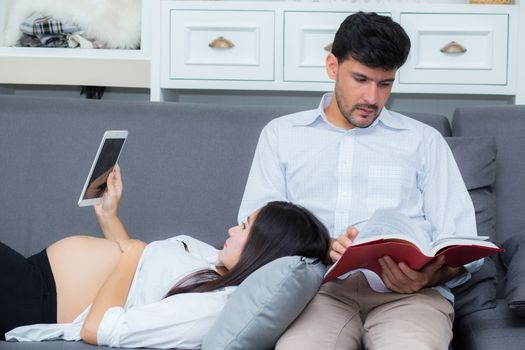 Asian couple family using a tablet at home, the young mother is pregnant lying on sofa looking tablet and husband reading book.