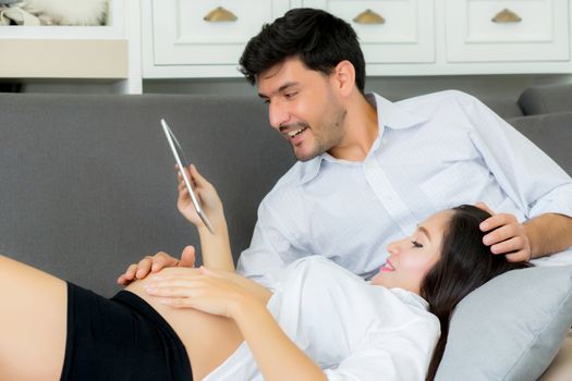 Asian couple family young mother and husband using a tablet at home, woman and man is pregnant lying on sofa looking tablet read book.