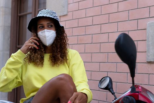 Portrait of an attractive young black woman sitting and talking with the mobile phone outdoors with her mask off. Concept communication