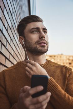 Smiling man listening to music on headphones and leaning against brick wall.