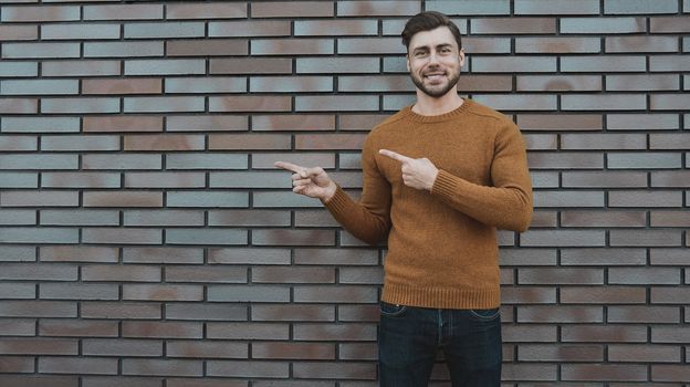 The horizontal look of an attractive young man who advertises something new, points his finger to the side, stands against a brick wall. People and lifestyle