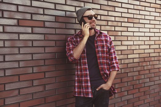 Hipster calling in city street on brick wall background. Amazing man holding smartphone in smart casual wear standing. Urban young professional lifestyle.