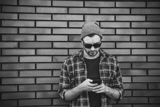 Hipster sms texting phone app in city street on brick wall background. Amazing man holding smartphone in smart casual wear standing.