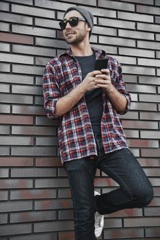 Hipster sms texting phone app in city street on brick wall background. Amazing man holding smartphone in smart casual wear standing.