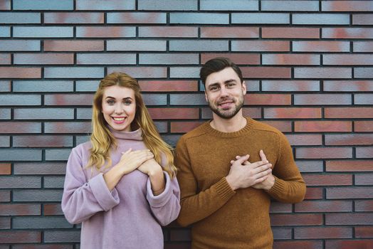 Graceful cheerful positive young woman and man have satisfied expressions, keep both hands on heart, feel thankful to guests, being in high spirit, stand against white background.