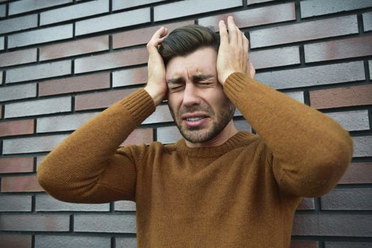 Man feeling painful emotions, screaming, witnessing death of close friend, being miserable and devastated, holding palms on head, yelling from pain and stress, standing unhappy over brown brick wall.