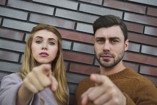 Hey you. Portrait of serious bearded man and woman in casual style standing and pointing at camera with serious face. outdoor shoot, isolated on brick wall background.