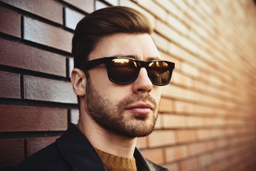 Portrait of stylish handsome young man with bristle standing outdoors and leaning on brick wall.