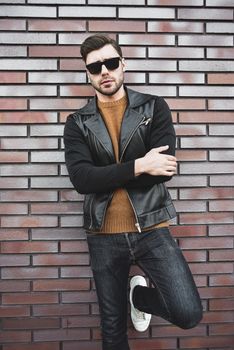 Portrait of stylish handsome young man with bristle standing outdoors and leaning on brick wall.