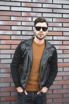 Portrait of stylish handsome young man with bristle standing outdoors and leaning on brick wall.