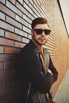 Portrait of stylish handsome young man with bristle standing outdoors and leaning on brick wall.