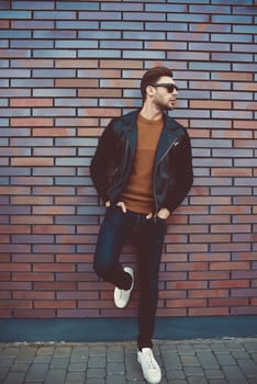 Portrait of stylish handsome young man with bristle standing outdoors and leaning on brick wall.