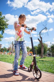 A child in torn jeans rides a scooter in a summer park. Modern childhood