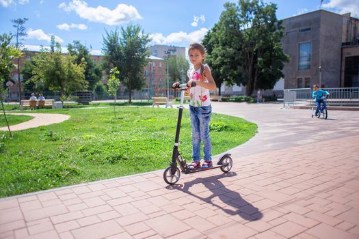 A child in torn jeans rides a scooter in a summer park. Modern childhood