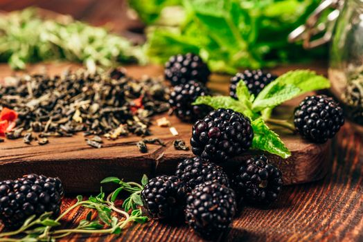 Heap of Dry Green Tea and Fresh Blackberries on Wooden Cutting Board. Bundles of Mint and Thyme Leaves.