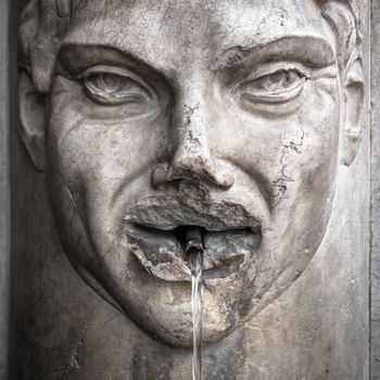 Detail of an old Italian fountain whose jet of water flows from the mouth of a face carved in marble.