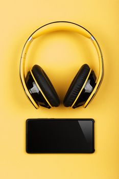 Close up modern wireless plastic yellow headphones with big cushions and black mobile phone on table, elevated top view, directly above