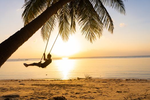 The guy enjoys the sunset riding on a swing on the ptropical beach. Silhouettes of a guy on a swing hanging on a palm tree, watching the sunset in the water