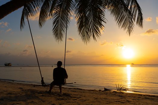 The guy enjoys the sunset riding on a swing on the ptropical beach. Silhouettes of a guy on a swing hanging on a palm tree, watching the sunset in the water