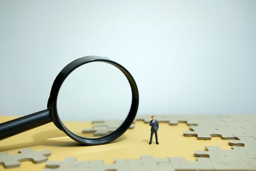 Business strategy conceptual photo - Miniature businessman standing behind magnifier glass in the middle of jigsaw puzzle