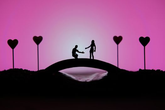 miniature people / toy photography - conceptual valentine holiday illustration. A man proposing a girl silhouette above the bridge with heart lamp
