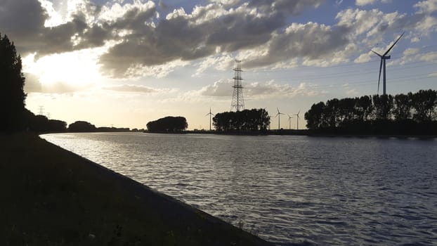 Sunset silhouette on the shore of a canal in industrial area, wind turbine and electricity pylon in the distance