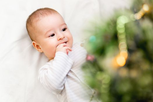 Baby boy or girl lying under Christmas tree. Little child plays with fir tree decorated for New Year celebration. Cute kid portrait. Winter holiday spirit.