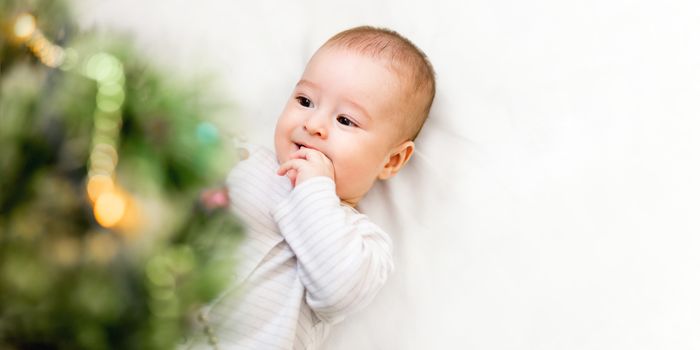 Baby boy or girl lying under Christmas tree. Little child plays with fir tree decorated for New Year celebration. Cute kid portrait. Winter holiday spirit. Banner with copy space.