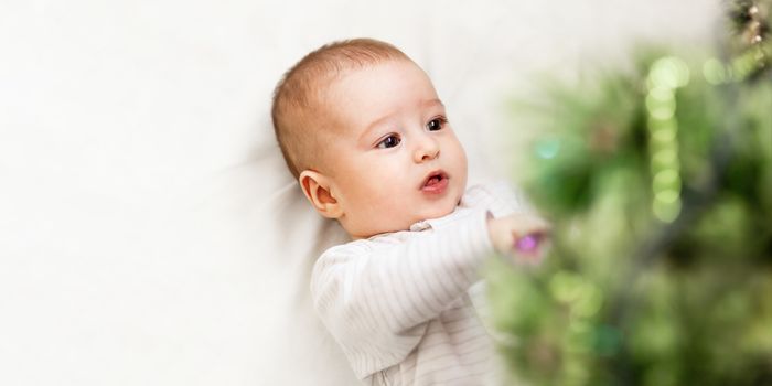 Baby boy or girl lying under Christmas tree. Little child plays with fir tree decorated for New Year celebration. Cute kid portrait. Winter holiday spirit. Banner with copy space.