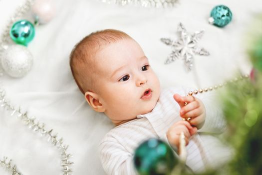 Baby boy or girl lying under Christmas tree. Little child plays with fir tree decorated for New Year celebration. Cute kid portrait. Winter holiday spirit.