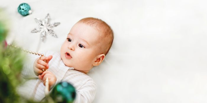 Baby boy or girl lying under Christmas tree. Little child plays with fir tree decorated for New Year celebration. Cute kid portrait. Winter holiday spirit. Banner with copy space.