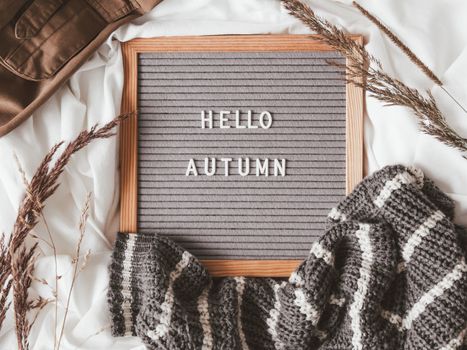 Top view on letter board with season greeting HELLO AUTUMN. Crumpled textile background with dried grass, grey knitted sweater and brown warm outfits.