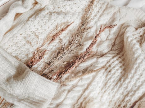 White knitted sweater with dried grass. Autumn concept flat lay. Warm cardigan with plants on it. Rustic authentic lifestyle background.