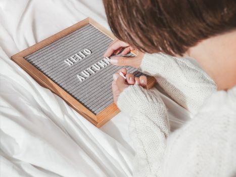 Woman in white knitted sweater typing HELLO AUTUMN on letter board. Crumpled textile background. Season greeting concept. Sunny, warm and cozy home.