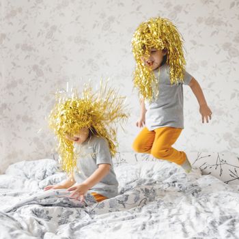 Twin brothers in bright yellow wigs jump on bed. Morning fun in cozy home. Funny children in colorful holiday wigs. Photocollage.