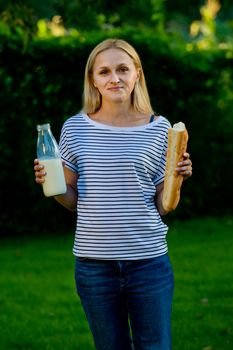 Portrait of a young woman who stands in her garden and holds a glass bottle with milk and a baguette.