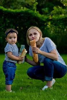 A young woman in jeans and a striped T-shirt gives her daughter milk from a glass bottle. Family picnic.