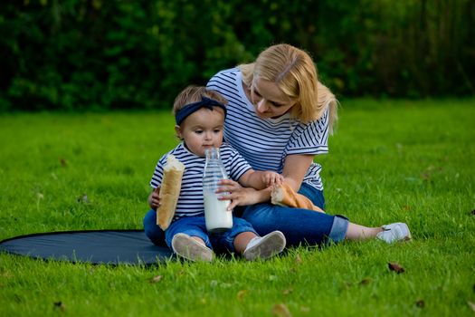 A young woman in jeans and a striped T-shirt gives her daughter milk from a glass bottle. Family picnic.