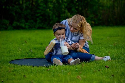 A young woman in jeans and a striped T-shirt gives her daughter milk from a glass bottle. Family picnic.