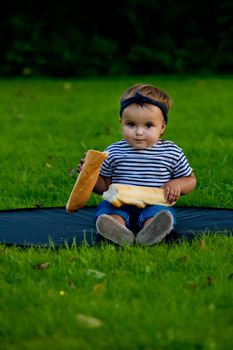 A little pretty baby girl sits on the lawn in the garden and holds a fresh baguette.