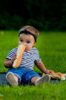 A little pretty baby girl sits on the lawn in the garden and holds a fresh baguette.