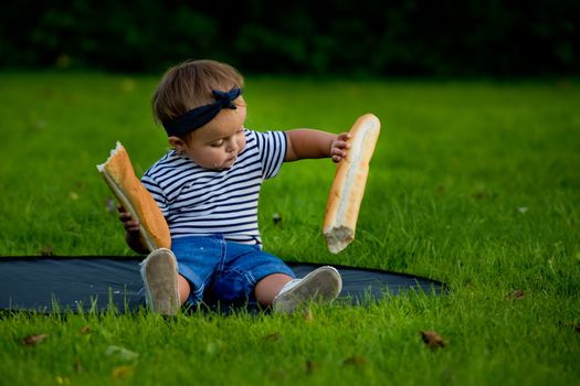 A little pretty baby girl sits on the lawn in the garden and holds a fresh baguette.
