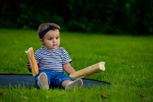 A little pretty baby girl sits on the lawn in the garden and holds a fresh baguette.