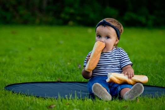 A little pretty baby girl sits on the lawn in the garden and holds a fresh baguette.