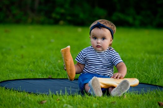 A little pretty baby girl sits on the lawn in the garden and holds a fresh baguette.