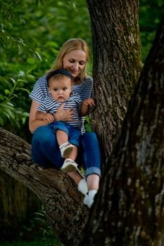 A young woman in jeans and a striped T-shirt and her little daughter are sitting on a tree.