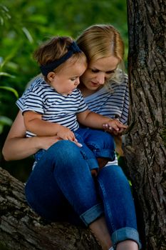 A young woman in jeans and a striped T-shirt and her little daughter are sitting on a tree.