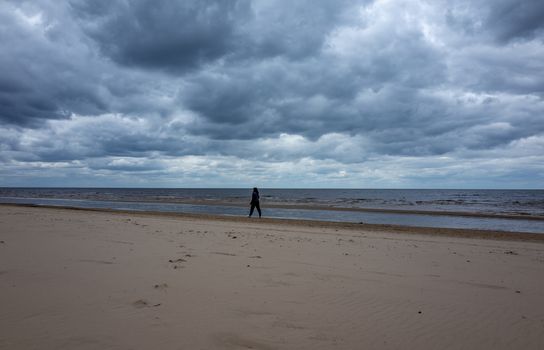 A girl in a warm black jacket walks along the seashore on a sandy beach in cloudy, cool weather.