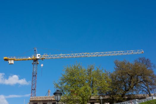 Long yellow arrow of a construction crane against the background of a clear sky and green trees
